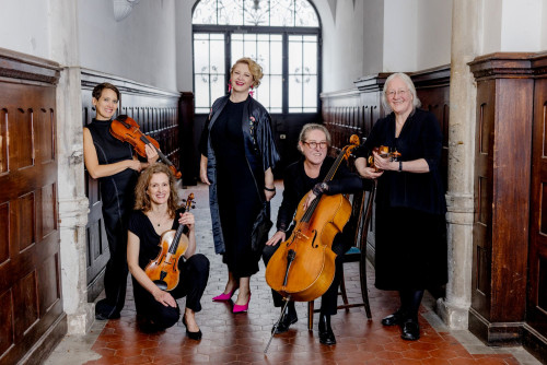 Fünf Frauen stehen und sitzen in einem holzgetäfelten Flur und halten Streichinstrumente, darunter Geigen, eine Bratsche und ein Cello. Sie lächeln und sind schwarz gekleidet, eine Frau steht in der Mitte und trägt ein Kleid und rosa Schuhe.