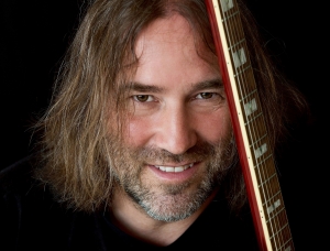 Ein Mann mit langen braunen Haaren und Bart lächelt in die Kamera und hält vor einem dunklen Hintergrund eine Gitarre senkrecht neben seinem Gesicht.