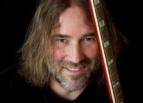 Ein Mann mit langen braunen Haaren und Bart lächelt in die Kamera und hält vor einem dunklen Hintergrund eine Gitarre senkrecht neben seinem Gesicht.