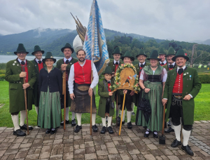 Eine Gruppe von Menschen in traditioneller bayerischer Kleidung posiert im Freien auf einem Steinweg, mit grünen Bergen und einem See im Hintergrund. Einige halten Fahnen und eine verzierte Holztafel in der Hand. Die Stimmung ist festlich und fröhlich.