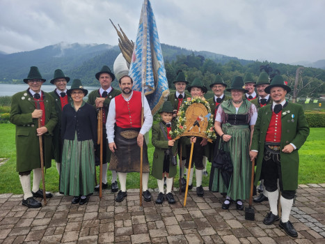 Eine Gruppe von Menschen in traditioneller bayerischer Kleidung posiert im Freien auf einem Steinweg, mit grünen Bergen und einem See im Hintergrund. Einige halten Fahnen und eine verzierte Holztafel in der Hand. Die Stimmung ist festlich und fröhlich.
