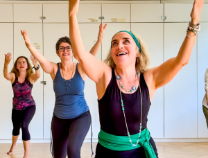 Drei Frauen in Trainingskleidung heben freudig die Arme, während sie in einem hellen Raum mit Holzfußboden und weißen Schränken an einem Gruppentanz- oder Fitnesskurs teilnehmen.