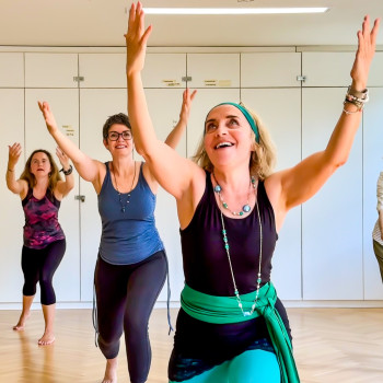 Drei Frauen in Trainingskleidung heben freudig die Arme, während sie in einem hellen Raum mit Holzfußboden und weißen Schränken an einem Gruppentanz- oder Fitnesskurs teilnehmen.