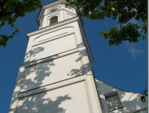 Ein Kirchturm der alten Sendlinger Kirche von unten gesehen, umrahmt von grünen Zweigen und Blättern, mit Schattenwurf auf dem Gebäude und einem blauen Himmel mit ein paar Wolken im Hintergrund.