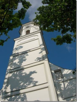 Ein Kirchturm der alten Sendlinger Kirche von unten gesehen, umrahmt von grünen Zweigen und Blättern, mit Schattenwurf auf dem Gebäude und einem blauen Himmel mit ein paar Wolken im Hintergrund.