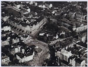 Eine Schwarz-Weiß-Luftaufnahme einer Stadtkreuzung mit mehreren zusammenlaufenden Straßen, umgeben von historischen mehrstöckigen Gebäuden, Grünflächen und Bäumen. Die Szene scheint aus dem frühen 20. Jahrhundert zu stammen.