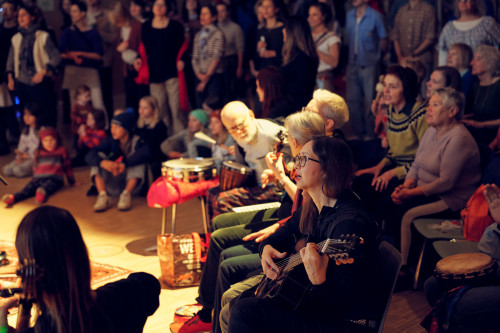 Eine Gruppe von Menschen versammelt sich in einem Raum, einige sitzend, andere stehend, und hört Live-Musik. Mehrere Personen spielen Instrumente wie Gitarre und Schlagzeug. Das Publikum scheint engagiert und aufmerksam zu sein.