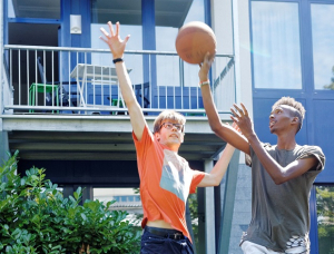Zwei junge Leute spielen im Freien Basketball; der eine im orangefarbenen Hemd versucht, den anderen im grauen Hemd zu blocken, der den Ball in der Nähe des Korbes hält, mit einem Gebäude und Grün im Hintergrund.
