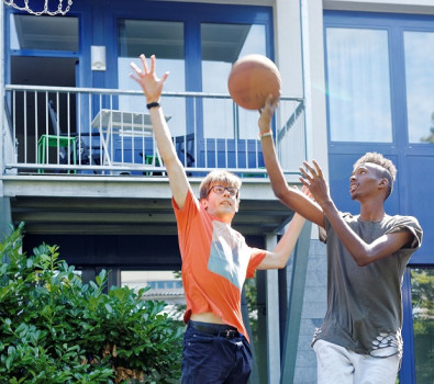 Zwei junge Leute spielen im Freien Basketball; der eine im orangefarbenen Hemd versucht, den anderen im grauen Hemd zu blocken, der den Ball in der Nähe des Korbes hält, mit einem Gebäude und Grün im Hintergrund.