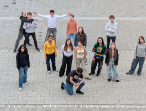 Eine Gruppe von 15 jungen Menschen steht auf einer gepflasterten Fläche, die in Form des Buchstabens 
