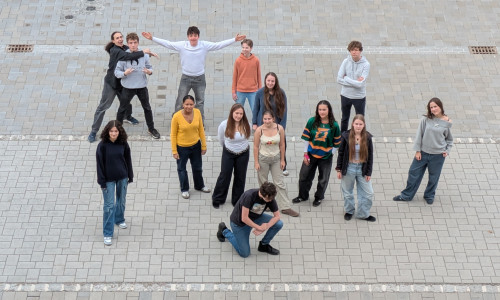 Eine Gruppe von 15 jungen Menschen steht auf einer gepflasterten Fläche, die in Form des Buchstabens 