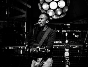 Ein Mann spielt Akustikgitarre und singt in ein Mikrofon auf der Bühne. Die Szene ist schwach beleuchtet, im Hintergrund leuchtet eine runde Leuchte und schafft eine stimmungsvolle, intime Atmosphäre.