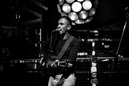 Ein Mann spielt Akustikgitarre und singt in ein Mikrofon auf der Bühne. Die Szene ist schwach beleuchtet, im Hintergrund leuchtet eine runde Leuchte und schafft eine stimmungsvolle, intime Atmosphäre.