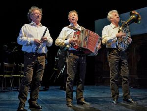 Drei ältere Männer stehen in passenden weißen Hemden und dunklen Hosen auf der Bühne und spielen jeweils ein Instrument: Klarinette, Akkordeon und Horn. Sie spielen im Scheinwerferlicht auf der Bühne.