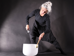 Eine ältere Frau mit grauem Haar in einem Dutt, schwarz gekleidet, hält ein Mikrofon in der Hand und schlägt eine große weiße Klangschale mit einem Schlegel an. Sie steht in einer dramatischen Pose vor einem dunklen Hintergrund.