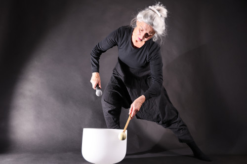 Eine ältere Frau mit grauem Haar in einem Dutt, schwarz gekleidet, hält ein Mikrofon in der Hand und schlägt eine große weiße Klangschale mit einem Schlegel an. Sie steht in einer dramatischen Pose vor einem dunklen Hintergrund.