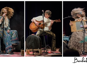 zwei Frauen und ein Mann spielen mit ihren Instrumenten sitzend auf einer Bühne. Der Mann spielt eine Gitarre, eine der Frauen spielt eine Zitter