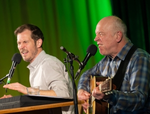 Zwei Männer treten auf der Bühne auf; einer singt leidenschaftlich in ein Mikrofon, während er Akustikgitarre spielt, der andere gestikuliert ausdrucksstark, während er in ein Mikrofon spricht oder singt. Beide wirken lebhaft und engagiert.