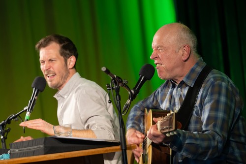 Zwei Männer treten auf der Bühne auf; einer singt leidenschaftlich in ein Mikrofon, während er Akustikgitarre spielt, der andere gestikuliert ausdrucksstark, während er in ein Mikrofon spricht oder singt. Beide wirken lebhaft und engagiert.