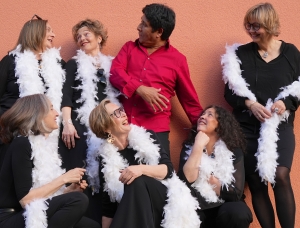Sechs Frauen in schwarzen Outfits und weißen Federboas posieren fröhlich um einen Mann im roten Hemd, der vor einer pfirsichfarbenen Wand steht. Alle lächeln und interagieren spielerisch.