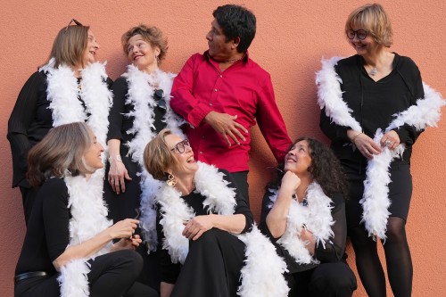 Sechs Frauen in schwarzen Outfits und weißen Federboas posieren fröhlich um einen Mann im roten Hemd, der vor einer pfirsichfarbenen Wand steht. Alle lächeln und interagieren spielerisch.