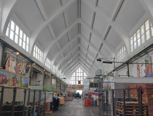 Blick in eine der geräumigen, überdachten Markthallen mit hoher, gewölbter weißer Decke, eingezäunten Ständen auf beiden Seiten, verschiedenen Waren und Erzeugnissen und dem freien Mittelgang