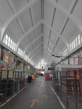 Blick in eine der geräumigen, überdachten Markthallen mit hoher, gewölbter weißer Decke, eingezäunten Ständen auf beiden Seiten, verschiedenen Waren und Erzeugnissen und dem freien Mittelgang