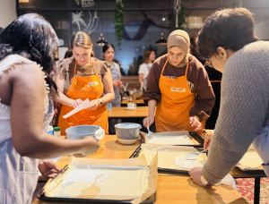 Vier Frauen in Schürzen stehen um einen Tisch herum und verteilen Teig auf mit Pergamentpapier ausgelegte Backbleche in einer hell erleuchteten Küche bei einem Kochkurs, während andere im Hintergrund zu sehen sind.