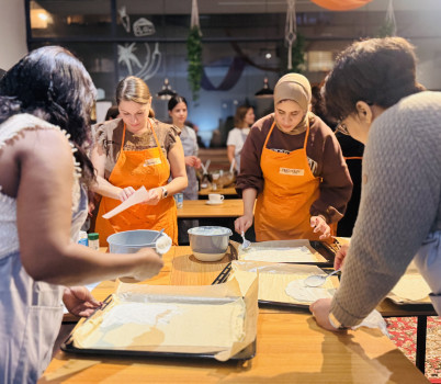 Vier Frauen in Schürzen stehen um einen Tisch herum und verteilen Teig auf mit Pergamentpapier ausgelegte Backbleche in einer hell erleuchteten Küche bei einem Kochkurs, während andere im Hintergrund zu sehen sind.