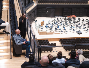 Eine Frau steht und gestikuliert, während sie zu einer Gruppe spricht, die in einem modernen Auditorium mit Blick auf eine leere Orchesterbühne mit vielen Stühlen und Notenständern sitzt.