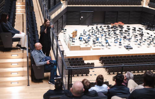 Eine Frau steht und gestikuliert, während sie zu einer Gruppe spricht, die in einem modernen Auditorium mit Blick auf eine leere Orchesterbühne mit vielen Stühlen und Notenständern sitzt.