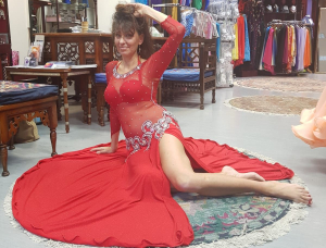 Eine Frau in einem durchsichtigen roten Bauchtanzkleid sitzt mit einem ausgestreckten und einem angewinkelten Bein auf einem runden Teppich und posiert anmutig in einem Tanzkostümgeschäft, umgeben von Kleidern und Holzmöbeln.