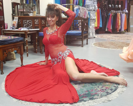 Eine Frau in einem durchsichtigen roten Bauchtanzkleid sitzt mit einem ausgestreckten und einem angewinkelten Bein auf einem runden Teppich und posiert anmutig in einem Tanzkostümgeschäft, umgeben von Kleidern und Holzmöbeln.