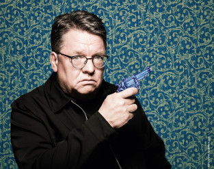 Ein ernst dreinblickender Mann mit Brille und schwarzer Jacke posiert vor einem gemusterten blauen und grünen Hintergrund und hält eine blaue Spielzeugpistole in der Hand.