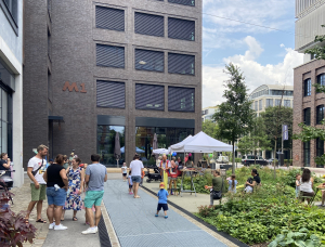 Menschen versammeln sich im Freien in der Nähe eines modernen Gebäudes mit großen Fenstern. Einige stehen oder sitzen, während andere weiße Pavillons besuchen. Der Bereich ist begrünt, und Kinder spielen auf dem Gehweg.