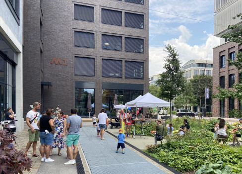 Menschen versammeln sich im Freien in der Nähe eines modernen Gebäudes mit großen Fenstern. Einige stehen oder sitzen, während andere weiße Pavillons besuchen. Der Bereich ist begrünt, und Kinder spielen auf dem Gehweg.
