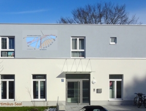 Ein modernes weißes Gebäude mit rechteckigen Fenstern und der Aufschrift „Bartimäus-Saal“. An der oberen Fassade ist ein Sonnenuhrdiagramm abgebildet. Zwei Fahrräder stehen in der Nähe des Eingangs.