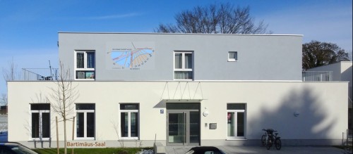 Ein modernes weißes Gebäude mit rechteckigen Fenstern und der Aufschrift „Bartimäus-Saal“. An der oberen Fassade ist ein Sonnenuhrdiagramm abgebildet. Zwei Fahrräder stehen in der Nähe des Eingangs.