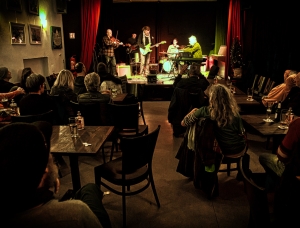 Eine kleine Band mit Gitarren, Keyboard und Geige spielt in einem schwach beleuchteten Saal auf der Bühne. Das Publikum sitzt an Tischen und schaut und hört zu, während warmes Licht eine intime Atmosphäre schafft.