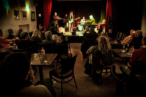 Eine kleine Band mit Gitarren, Keyboard und Geige spielt in einem schwach beleuchteten Saal auf der Bühne. Das Publikum sitzt an Tischen und schaut und hört zu, während warmes Licht eine intime Atmosphäre schafft.