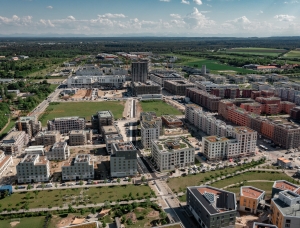 Luftaufnahme eines modernen Stadtgebiets mit mehreren mittelhohen Wohnhäusern, von denen sich einige im Bau befinden, umgeben von grünen Feldern, Straßen und vereinzelten Bäumen unter einem teilweise bewölkten Himmel.