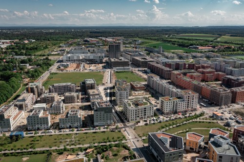 Luftaufnahme eines modernen Stadtgebiets mit mehreren mittelhohen Wohnhäusern, von denen sich einige im Bau befinden, umgeben von grünen Feldern, Straßen und vereinzelten Bäumen unter einem teilweise bewölkten Himmel.