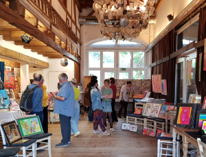 In einer rustikalen, gut beleuchteten Galerie mit Holzböden, freiliegenden Balken und einem großen Kronleuchter werden bunte Gemälde und Kunstwerke auf Staffeleien und Tischen ausgestellt. Mehrere Menschen sind in ein Gespräch vertieft.