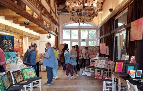 In einer rustikalen, gut beleuchteten Galerie mit Holzböden, freiliegenden Balken und einem großen Kronleuchter werden bunte Gemälde und Kunstwerke auf Staffeleien und Tischen ausgestellt. Mehrere Menschen sind in ein Gespräch vertieft.