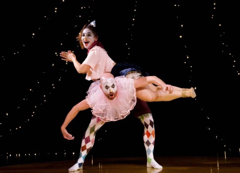 Zwei Clowns führen auf der Bühne eine akrobatische Nummer auf; ein Clown beugt sich nach vorne, während der andere seitlich auf dem Rücken balanciert. Beide tragen farbenfrohe Kostüme und Make-up, und im dunklen Hintergrund funkeln Lichterketten.