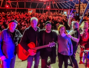 Eine fünfköpfige Band steht lächelnd mit Instrumenten auf der Bühne und posiert für ein Foto vor einer großen, jubelnden Menge unter einem Zelt mit bunter Beleuchtung.