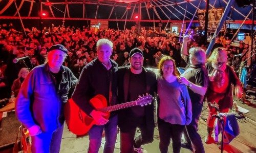 Eine fünfköpfige Band steht lächelnd mit Instrumenten auf der Bühne und posiert für ein Foto vor einer großen, jubelnden Menge unter einem Zelt mit bunter Beleuchtung.