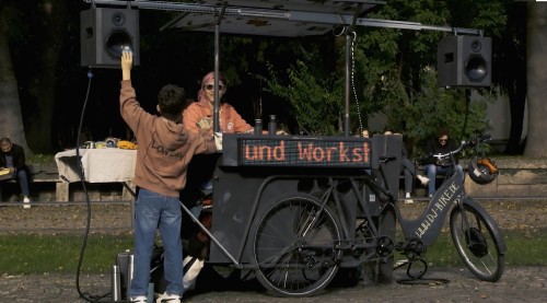 Eine Person in einem braunen Kapuzenpullover hebt die Hand neben einem mobilen DJ-Pult auf einem Fahrrad, wo eine weitere Person mit Kopfhörern Musik spielt. Auf dem Pult steht „und Works!“. Im Hintergrund sind Bäume und Menschen zu sehen.