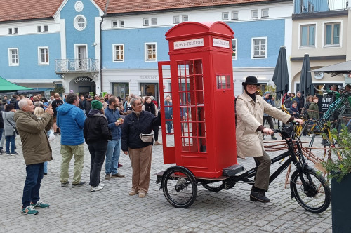 Eine Person im Anzug und mit Bowlerhut fährt auf einem Dreirad, an dem eine große rote Telefonzelle im britischen Stil befestigt ist, während eine Menschenmenge auf einem Stadtplatz zusieht und Fotos macht.