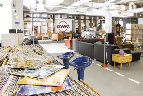 Ein moderner, geräumiger Raum mit Bücherregalen und Dekoartikeln, einem Loungebereich mit Sofas und einem Tisch mit Schallplatten im Vordergrund. Zwei blaue Barhocker stehen in der Nähe der Schallplatten.
